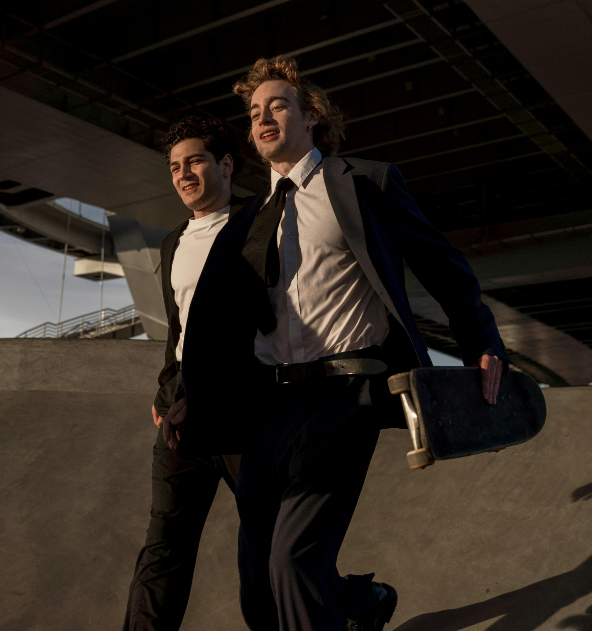 Two individuals in suits with one holding a skateboard, standing under a bridge.